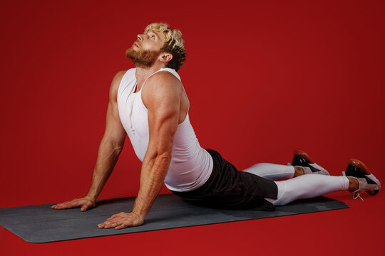 Full body side view young strong sporty toned sportsman man wear white clothes spend time in home gym lie on yoga mat do back stretch isolated on plain red background. Workout sport fit body concept.