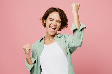 Young happy woman 20s she wear green shirt white t-shirt doing winner gesture celebrate clenching fists say yes isolated on plain pastel light pink background studio portrait People lifestyle concept