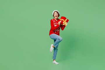 Fototapeta na wymiar Full body merry young woman wear knitted xmas sweater Santa hat posing hold red present box with gift ribbon bow isolated on plain pastel light green background. Happy New Year 2023 holiday concept.