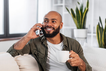 Mobile communication. Happy latin man talking on cellphone, sitting on couch and holding cup of coffee