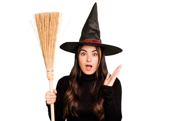 Young caucasian woman dressed as a witch holding a broom isolated on green chroma background surprised and shocked.