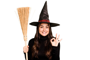 Young caucasian woman dressed as a witch holding a broom isolated on green chroma background cheerful and confident showing ok gesture.