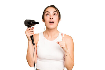 Young caucasian woman holding a hairdryer on green chroma background pointing upside with opened mouth.