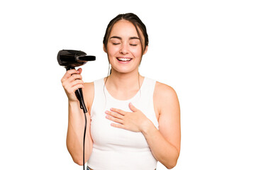 Young caucasian woman holding a hairdryer on green chroma background laughs out loudly keeping hand on chest.