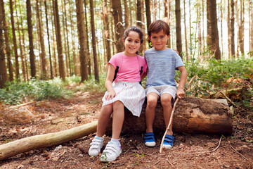Naklejka na ściany i meble Portrait Of Smiling Boy And Girl Walking In Summer Woodland Sitting On Log Wearing Backpacks