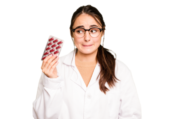 Young pharmacist woman holding a pills isolated on green chroma background confused, feels doubtful and unsure.