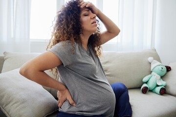 Pregnant woman sitting on the couch lower back pain and headache, strain on the spine during pregnancy. Lifestyle preparation for childbirth, last month of pregnancy