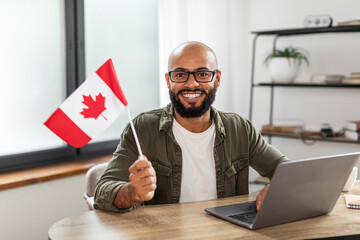 Happy latin man sitting with flag of Canada and using laptop computer, sitting at desk at home,...