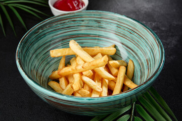 French fries in ceramic bowl on black concrete background. Composition with french fries and palm leaf on dark background. Junk food in modern style. Fried potatoes in asian style.