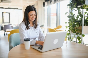 Fototapeta premium Freelance, business and e-commerce concept. Creative enthusiastic female journalist writing new article, typing on laptop, smiling, write-down project ideas, sit cafe, drink coffee working remote