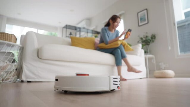 Internet of things smart tech urban house IOT appliances better life. Asia Gen Z people young woman easy relax neat chores enjoy home care to robot hoover clean floor mobile app at sofa living room.