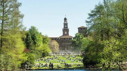 Palazzo del Arte and tourists on lawn in historical center of Milan on windy sunny day. Parch Sempion with lush greenery and river hyper lapse