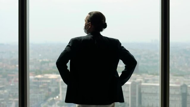 Business Man In A Suit And A Mask On His Face, Coming To The Window And Putting His Hands On His Hips. Then He Stands Looking Out Over The City From High Floor Of His Office, Turn Head