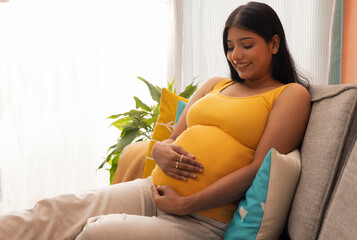 Smiling pregnant woman sitting on sofa at home with holding her abdomen