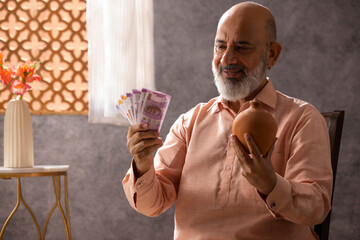 Portrait of happy senior man sitting at home with piggy bank and Indian currency
