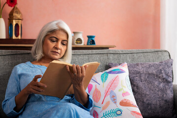 Old woman reading a book while sitting on sofa at home