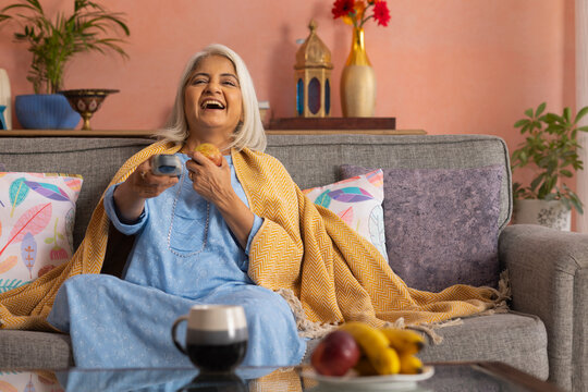 Cheerful old woman watching TV while sitting on sofa in living room