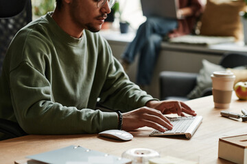 Close-up of young businessman in sweatshirt typing on computer keyboard while sitting by workplace...