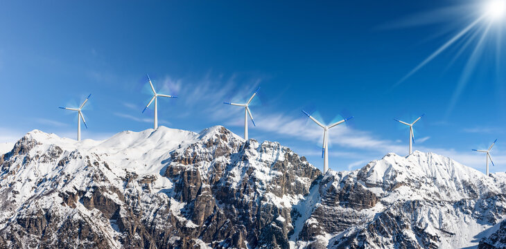 Group of white and blue wind turbines on the top of snow capped mountains against a clear blue sky with clouds and sunbeams.
