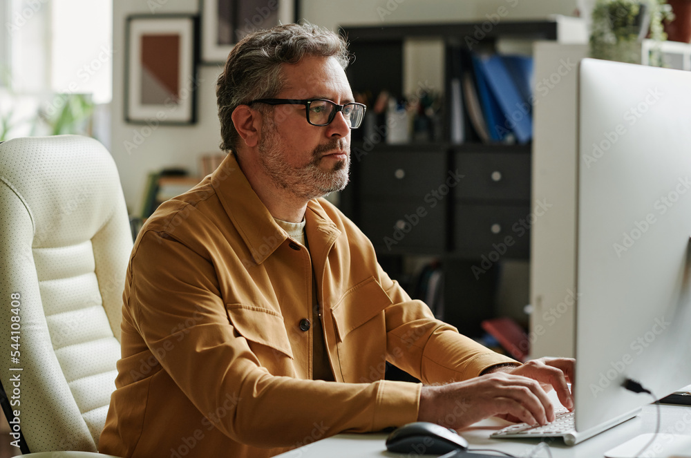 Leinwandbilder mature businessman with grey hair and beard typing on keyboard while sitting by workplace in front o