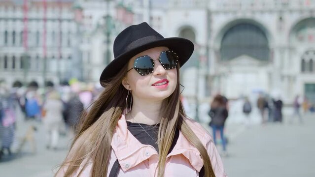 Stylish woman in black hat and sunglasses stands on St. Mark Square posing for photo. Happy long haired tourist enjoys vacation in Italy closeup