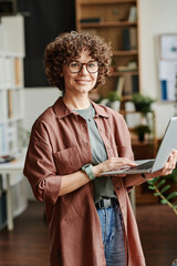 Young successful female designer with laptop standing in office and looking at camera while...