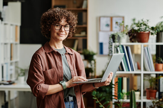 Young successful manager in casualwear standing in front of camera in coworking space and using laptop while analyzing online data