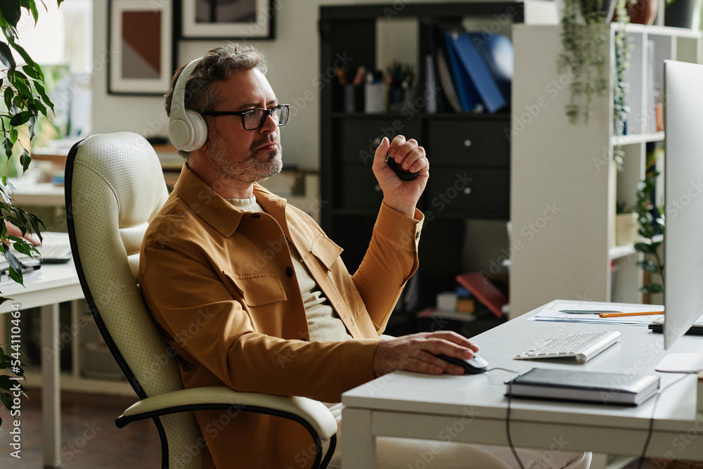 Poster Mature webdesigner squeezing antistress toy while sitting by workplace in front of computer monitor and clicking mouse during network