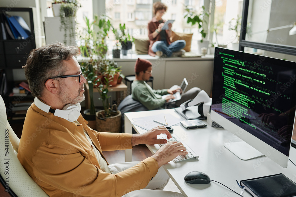 Poster Confident mature businessman looking at coded data on computer screen while sitting by workplace in front of monitor and typing
