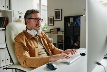 Serious grey haired programmer looking at computer screen while sitting by workplace in front of...