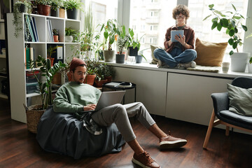 Young woman with tablet sitting on windowsill in front of male colleague with laptop networking in...