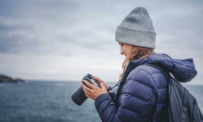 A young cheerful woman in a scarf jacket and a hat, a traveler professional photographer with a camera in her hands on the shore of the winter sea