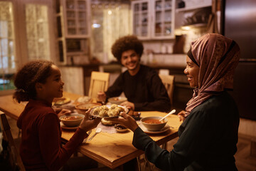 Happy Muslim mother and daughter passing food during family dinner at dining table.