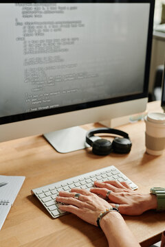 Young businesswoman or programmer pressing keys of computer keyboard while sitting by desk in front of monitor and decoding data