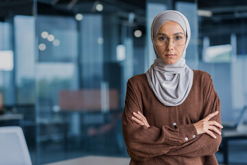 Serious and focused businesswoman in hijab and glasses looking at camera with crossed arms, working inside modern office building. - Powered by Adobe
