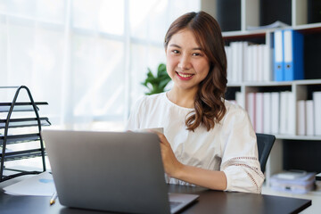 Beautiful Asian businesswoman sitting on her laptop happily working on her laptop and drinking coffee with a bright smile in the office and looking at the camera.
