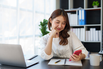 Obraz premium Smiling and charming Asian woman sit at her desk with a smartphone playing social media applications and transacting at her desk.