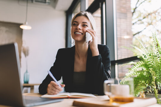 Cheerful female entrepreneur with laptop and textbook using cellphone device for calling and talking about business ideas, happy Caucasian woman discussing mobility communication while phoning