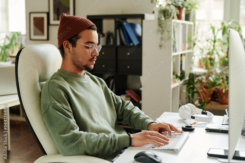 Poster Serious guy in casualwear typing on keyboard while sitting in front of computer monitor and working over new website or software