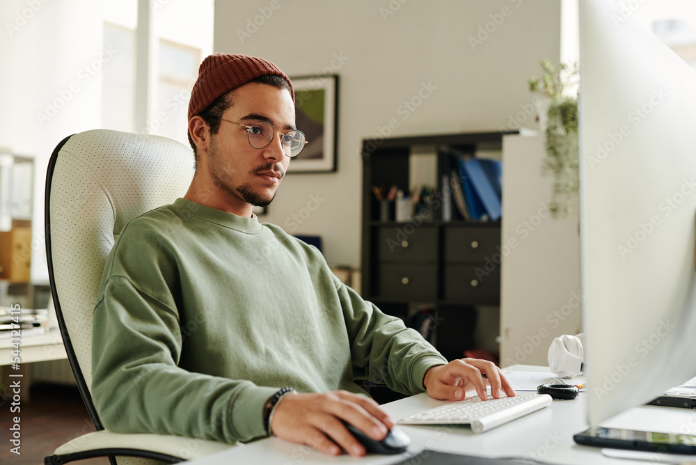 Leinwandbilder Professional IT engineer in eyeglasses looking at computer screen while keeping right hand on mouse and left one on keyboard