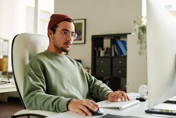 Professional IT engineer in eyeglasses looking at computer screen while keeping right hand on mouse and left one on keyboard