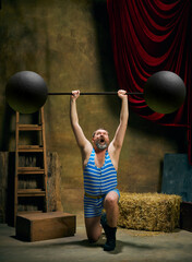 Vintage portrait of retro circus strongman wearing blue striped sports swimsuit training with...