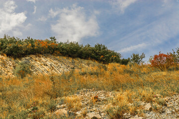 Beautiful picturesque yellow mountainside against a blue cloudy sky. The slope of the mountain against the sky. A rocky mountain slope with dry vegetation on a sunny autumn day.