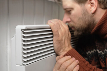 Young frozen man wearing a sweater shaking and freezing for winter cold with shock expression on face. Man is warming up hands with breath over electric heater. Discomfort spending time at home