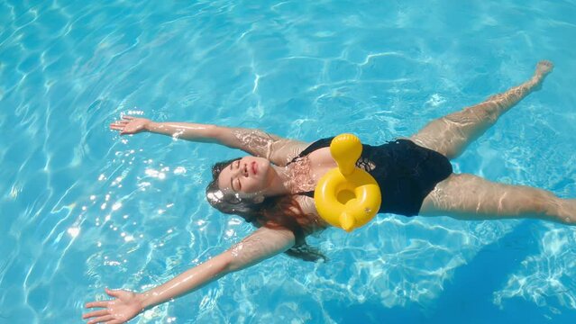 Cute girl in black bikini lying on blue water with yellow inflatable toy duck and smiling. Enjoy summer vacation in swimming pool with sun dogs on tropical resort at spa hotel. Female person bathing.