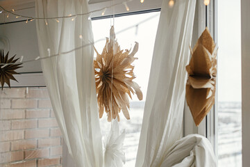 Stylish paper christmas stars and lights hanging in window in festive decorated boho room. Handmade paper swedish stars and garland, scandinavian decoration. Atmospheric christmas time