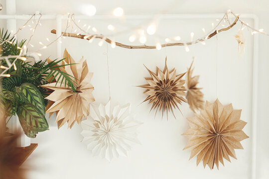 Stylish paper christmas stars and lights hanging on white wall background. Scandinavian festive decoration in boho room. Handmade paper swedish stars and garland. Atmospheric christmas time