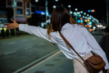 Asian business woman hail waving hand taxi on road in city street at night, Beautiful woman smiling using smartphone application hailing with hand up calling cab outdoor after late work, back view