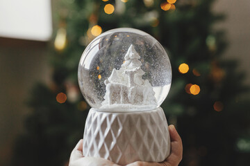 Stylish christmas snow globe in hand on background of christmas tree in lights in festive decorated...