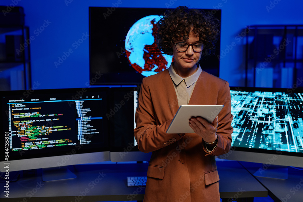 Poster Young serious female programmer using tablet while standing against computer monitors with coded data on screens in dark room